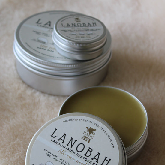 Two round silver metal tins of Lanobah Virgin Redensity Bar soap, one closed and one open showing the pale yellow-green bar inside, on a beige textured fabric background.