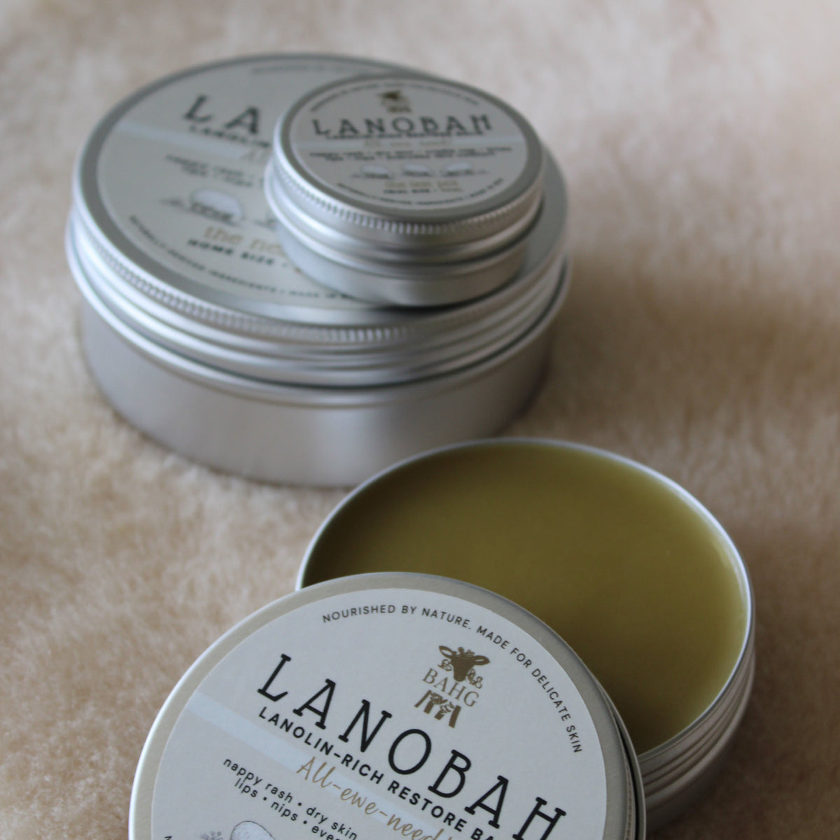 Two round silver metal tins of Lanobah Virgin Redensity Bar soap, one closed and one open showing the pale yellow-green bar inside, on a beige textured fabric background.