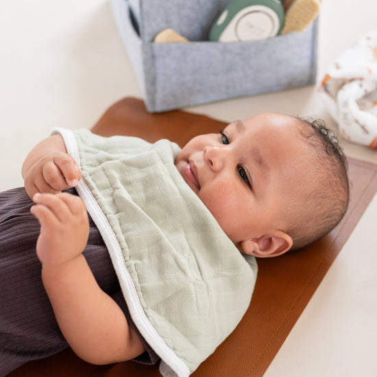Baby lying on a brown surface with a green bib, surrounded by toys and a storage bag.
