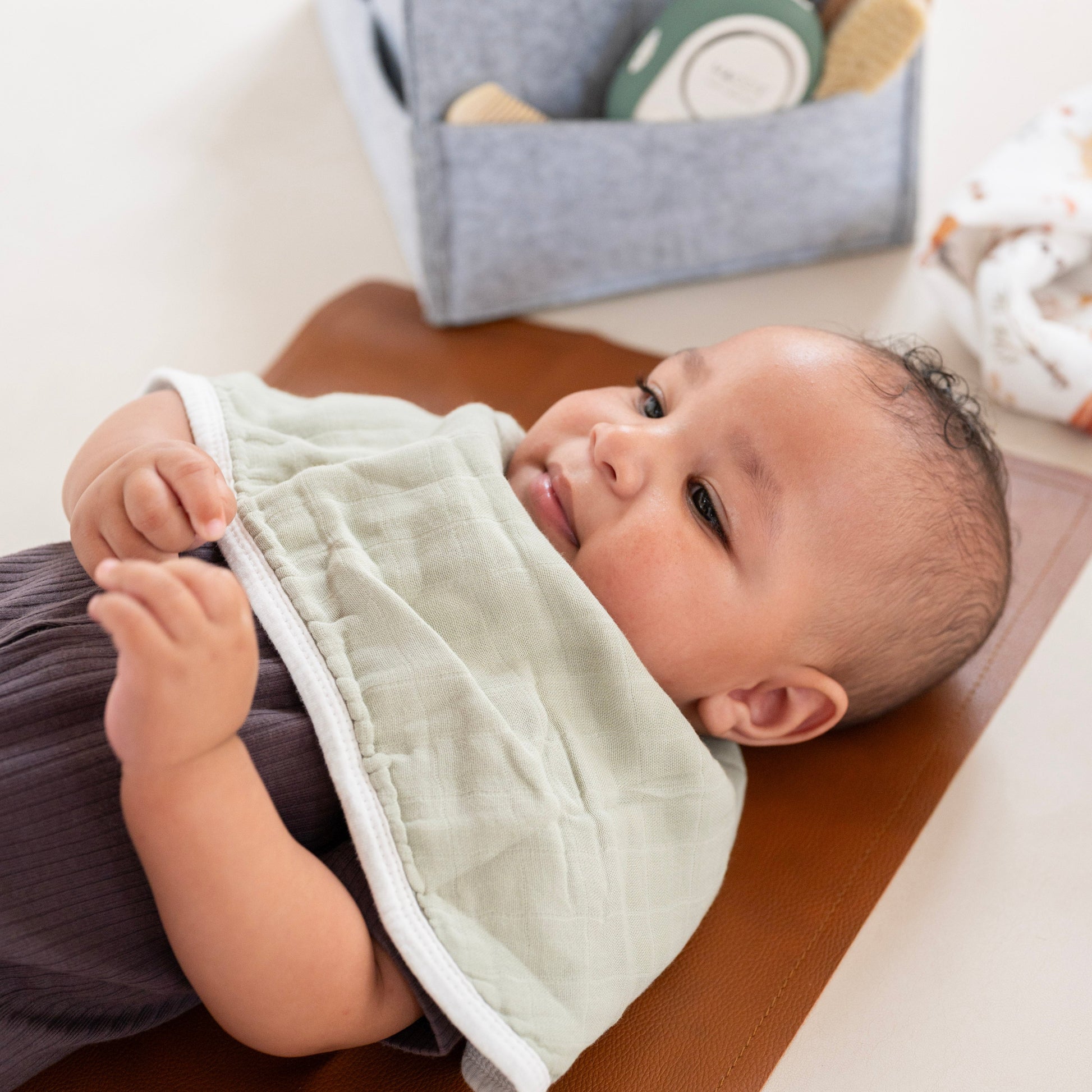 Baby lying on a brown surface with a green bib, surrounded by toys and a storage bag.