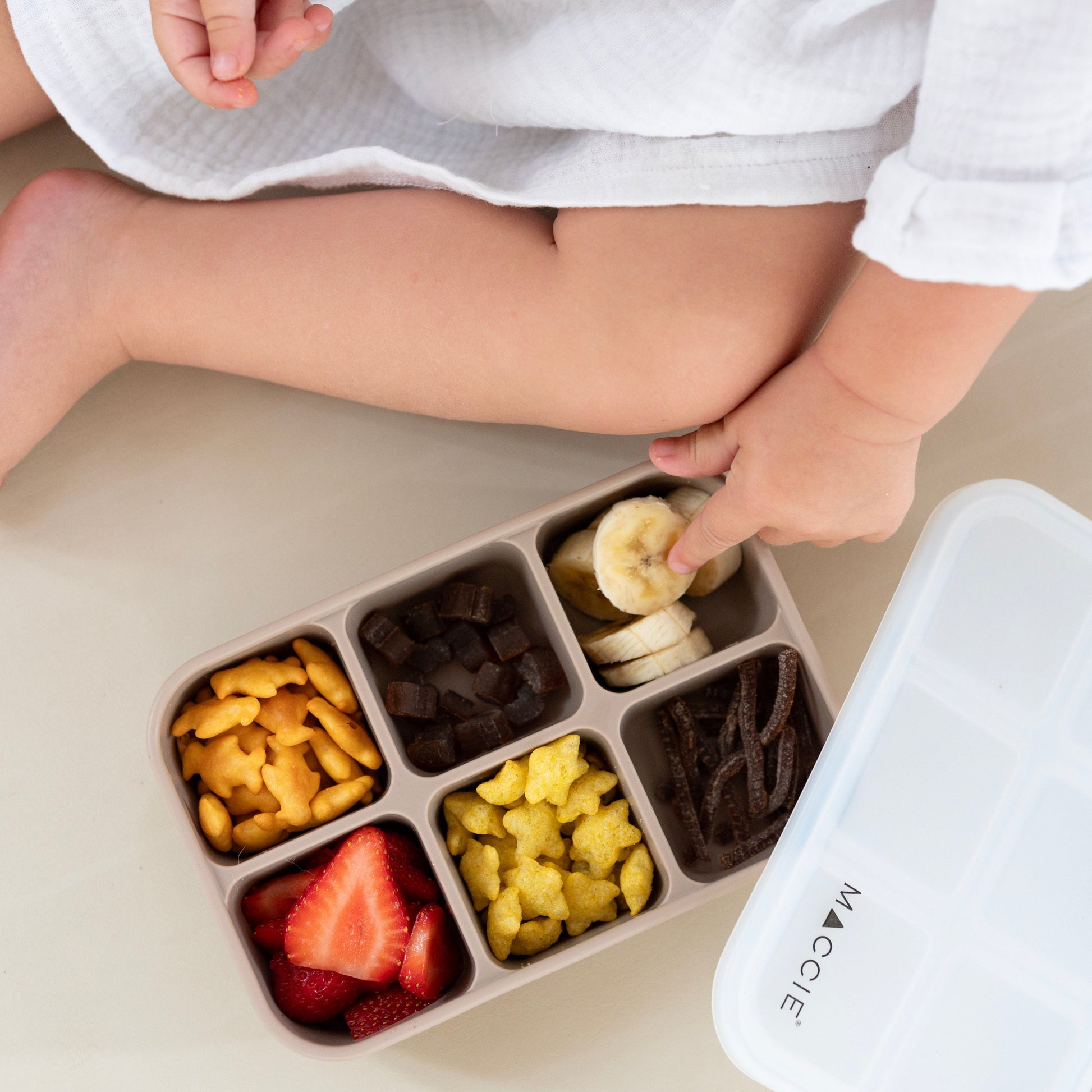 Child's hand reaching into a compartmentalized snack container with various foods on a neutral background.