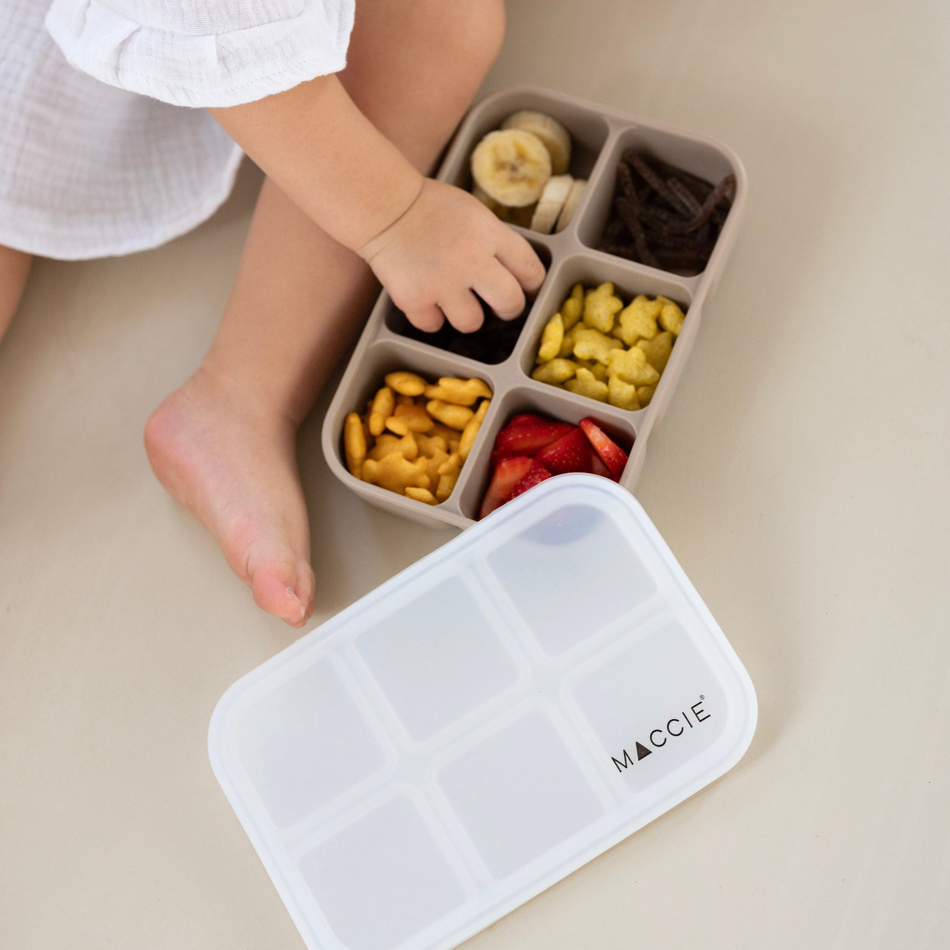 Child playing with a compartmentalized snack container on a light surface, with a MACKIE branded container in the foreground.