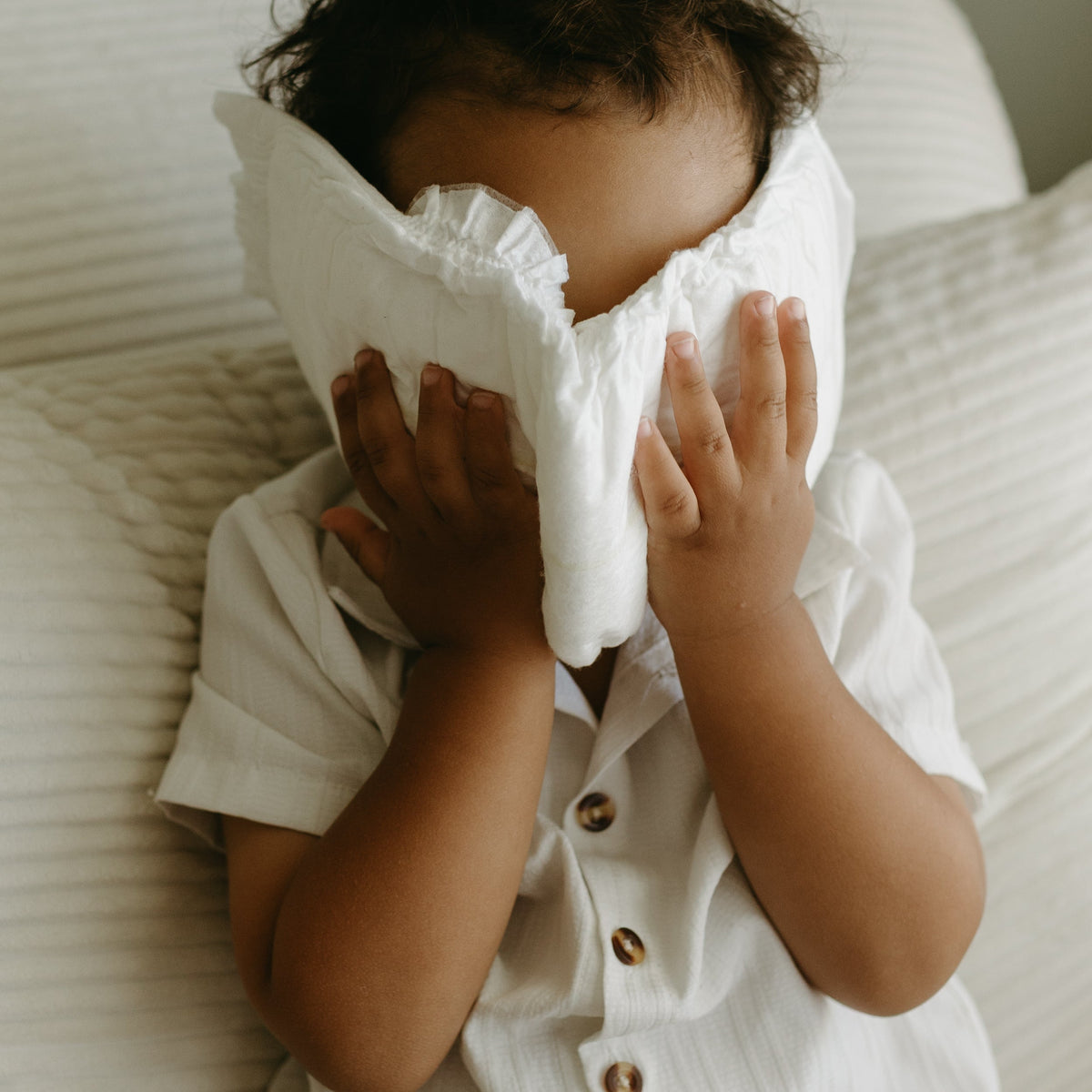 A baby wearing a white short-sleeve button-up shirt and a white diaper cover, sitting on a white ribbed sofa and covering their face with their hands.