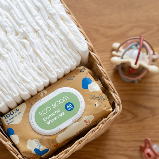A baby care gift set in a woven basket, featuring a pack of Eco Boom Bamboo Wipes, folded white diapers, and a wooden baby rattle on a wooden tabletop.