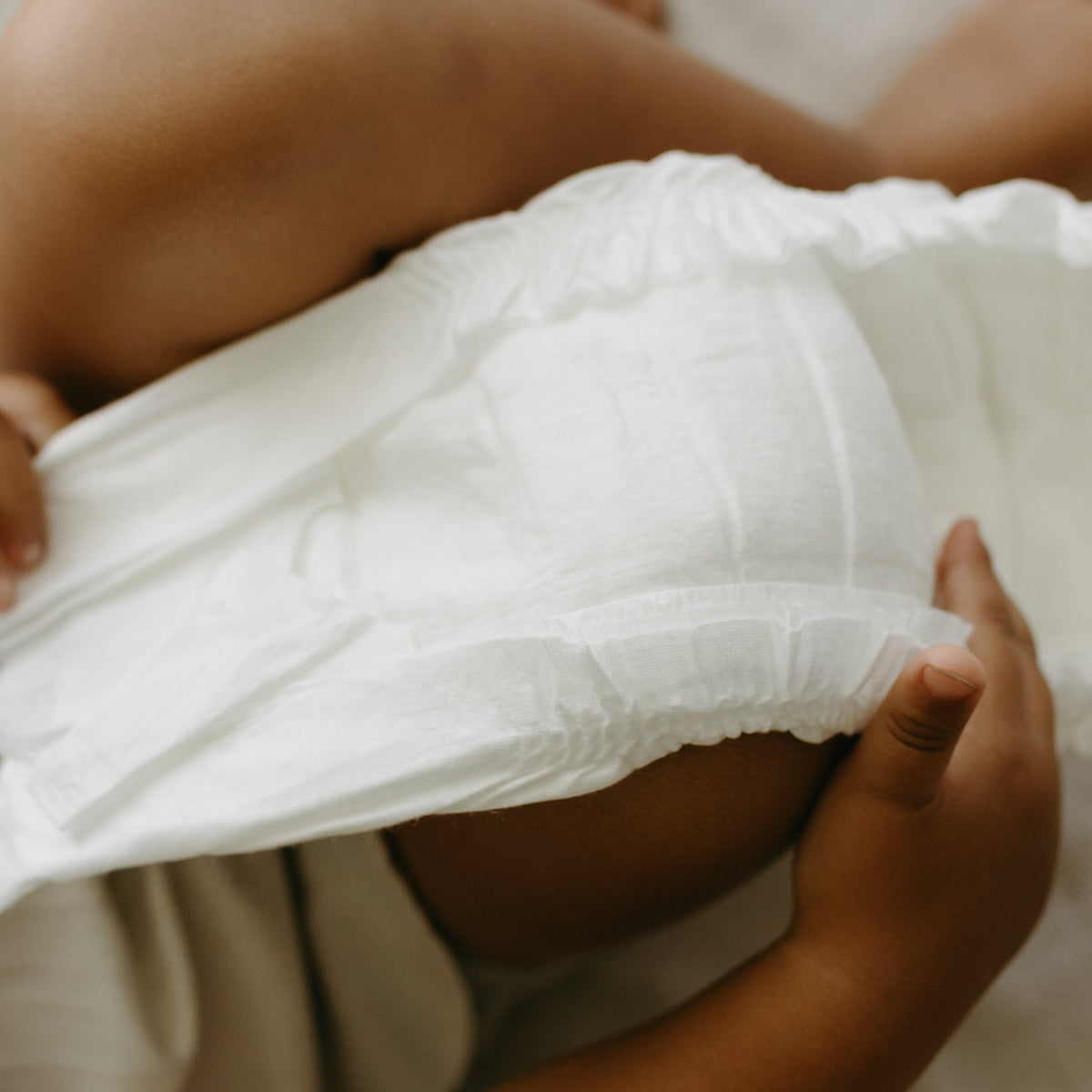 A close-up of a baby's legs and hands lifting a white disposable diaper, showing the soft texture of the diaper and the baby's skin against a light cream background.