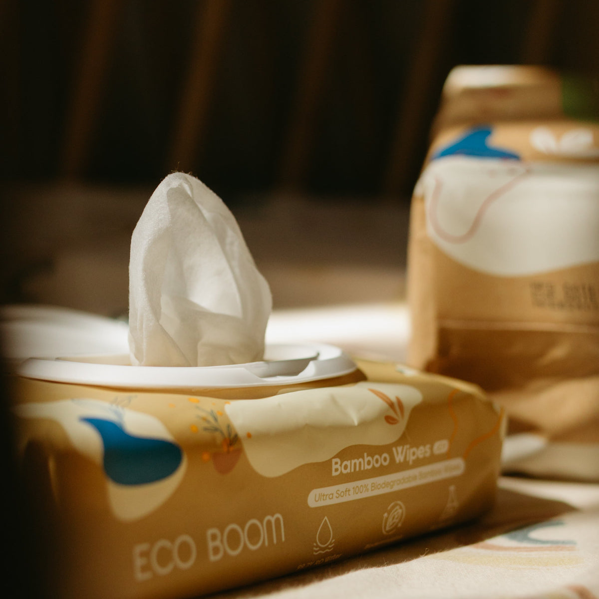 A close-up of a tissue box labeled ECO BOOM Bamboo Wipes with a white tissue pulled out, set in warm sunlight on a table with a blurred second box in the background.