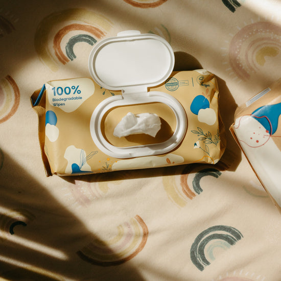 A package of baby wipes with the lid open, showing a single white wipe, placed on a patterned surface with warm sunlight and shadows.