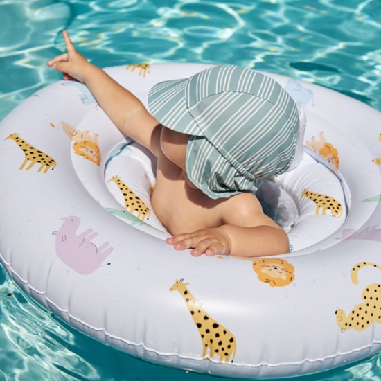 A baby wearing a striped sun hat sits in a pool float on a white inflatable ring with safari animal prints, floating in turquoise water.