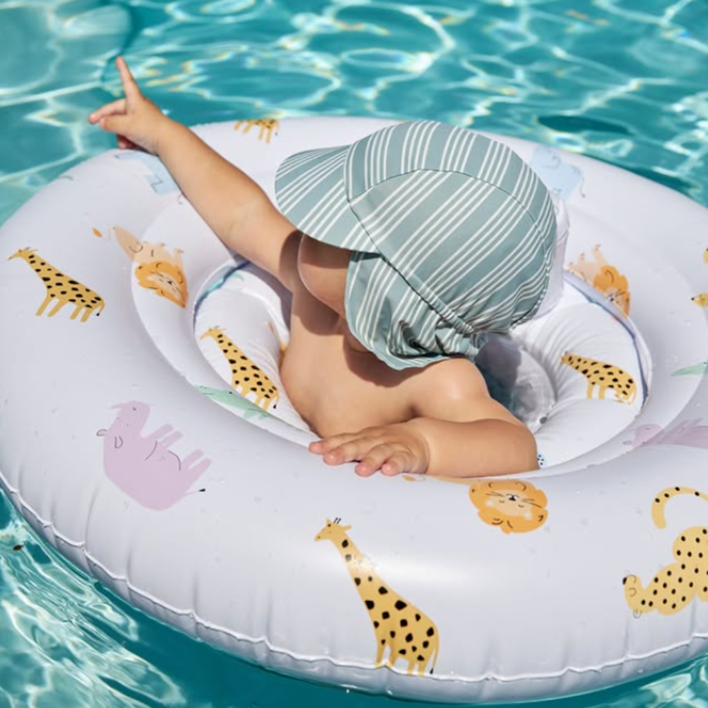 A baby wearing a striped sun hat sits in a pool float on a white inflatable ring with safari animal prints, floating in turquoise water.
