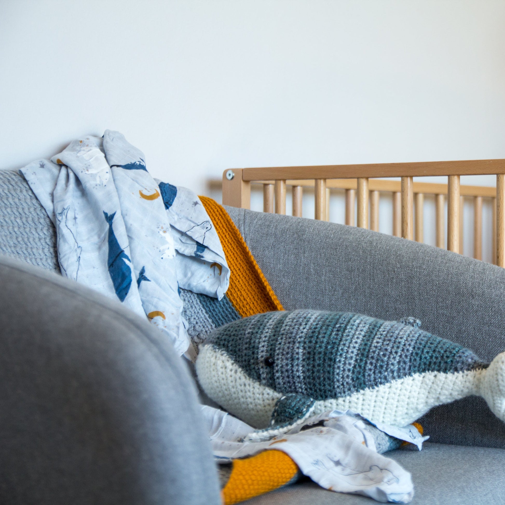Gray armchair with a knitted shark pillow and colorful blanket next to a wooden crib.