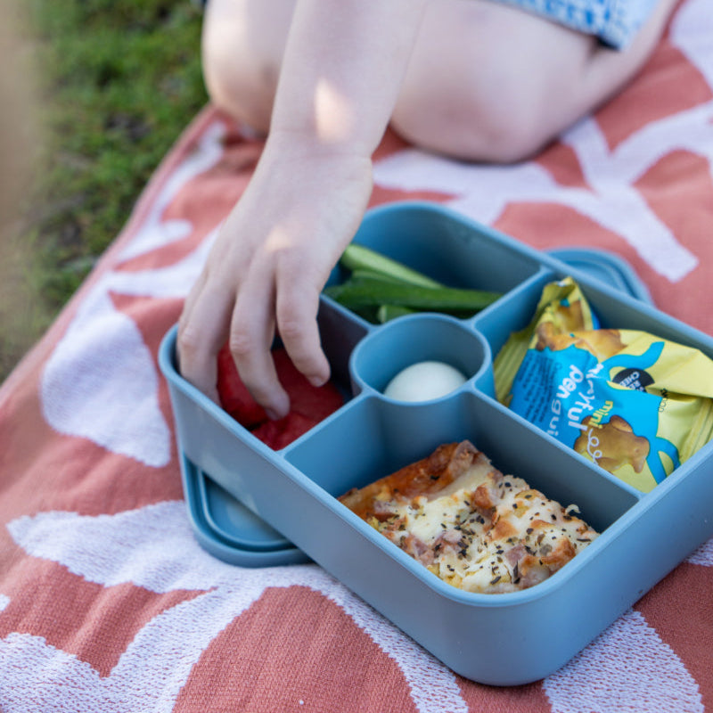 Person sitting on a blanket with a blue bento box containing food items.