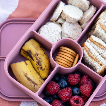 Pink bento box with compartments containing snacks like bananas, cookies, crackers, and berries on a patterned surface.