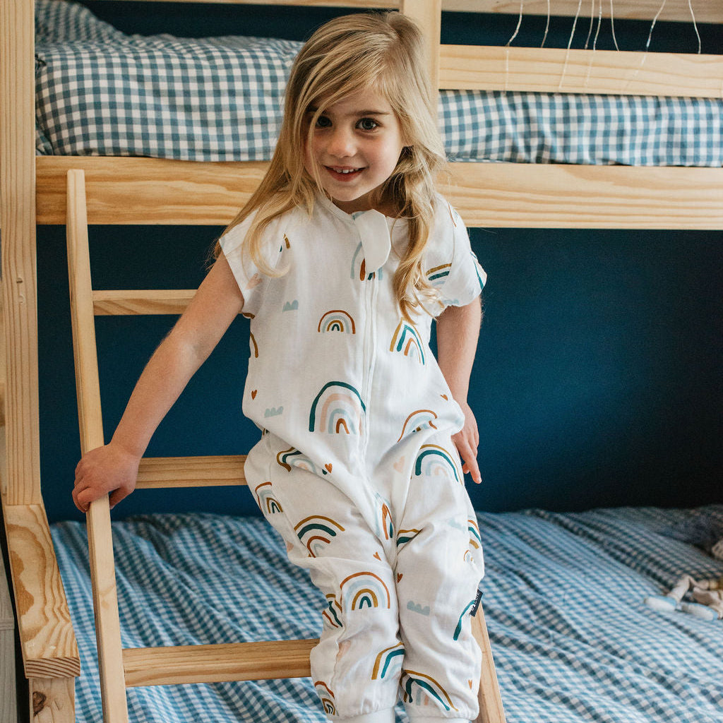 Child in a rainbow-patterned onesie standing on a wooden ladder of a bunk bed with blue checkered bedding.