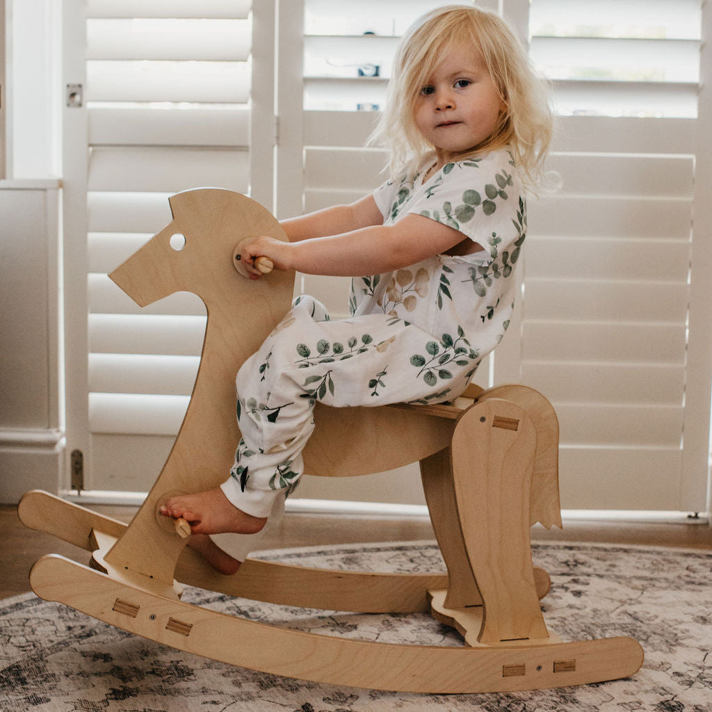Child sitting on a wooden rocking horse in a room with white shutters.