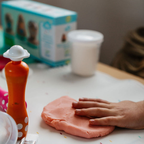 Pink play dough on a table with a hand touching it, surrounded by toys and a box.