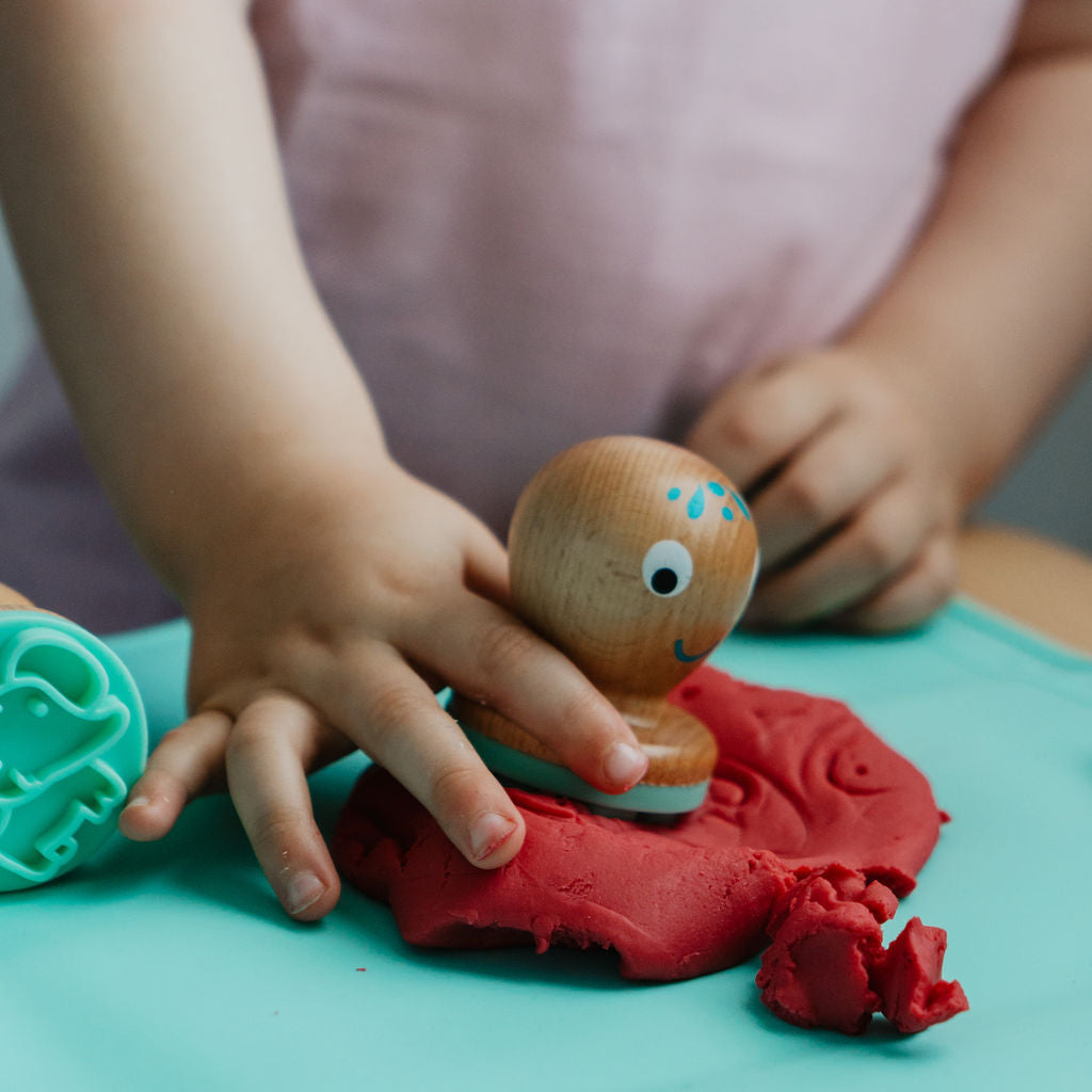 Child playing with red play dough and a wooden toy on a green surface