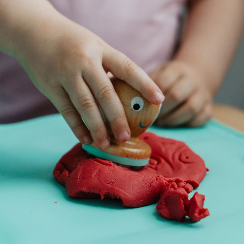 Child playing with a fish-shaped cookie cutter in red play dough on a light blue surface.