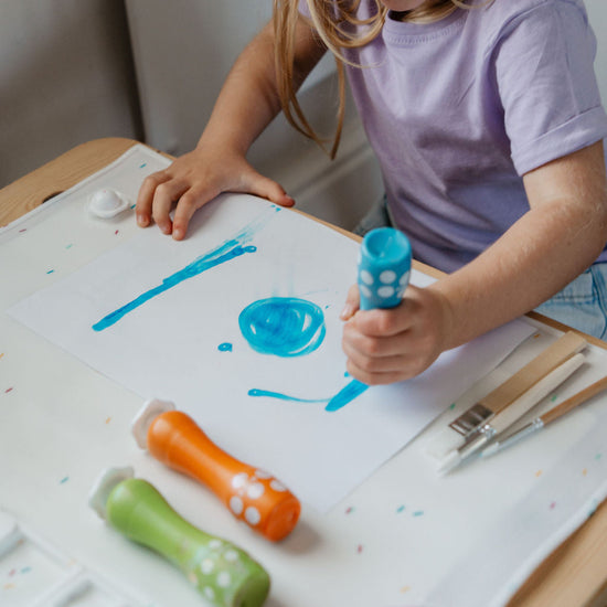 Child painting on a white board with blue paint and holding a green paintbrush.