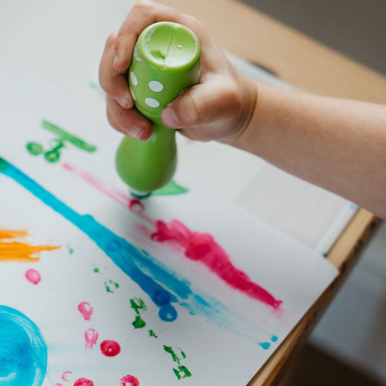 Child's hand holding a green marker with polka dots on a piece of paper with colorful paint splashes.