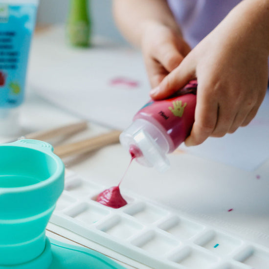 Person pouring a red liquid from a bottle into a white mold on a table with various items.