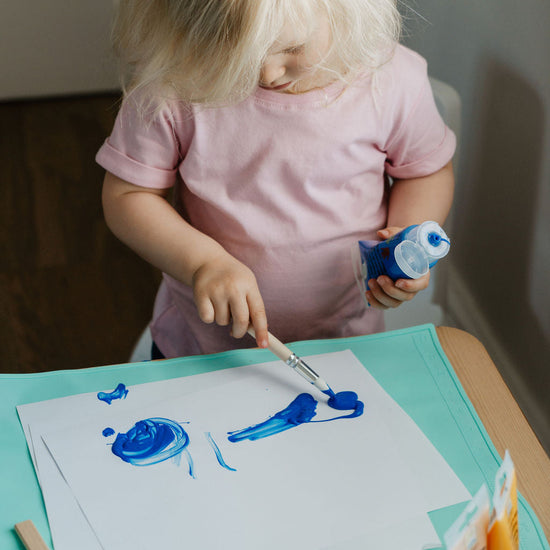 Child painting on paper with blue paint at a table.