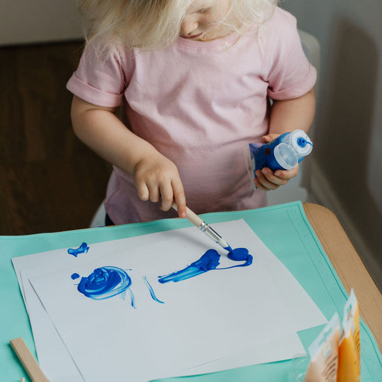 Child painting on paper with blue paint at a table.