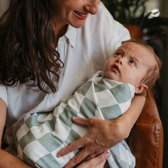 Woman holding a baby wrapped in a green and white checkered blanket indoors.