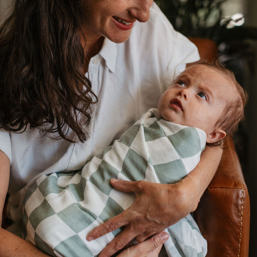 Woman holding a baby wrapped in a green and white checkered blanket indoors.
