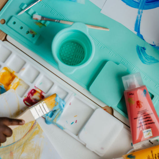 Children painting with brushes on a green art board with art supplies.