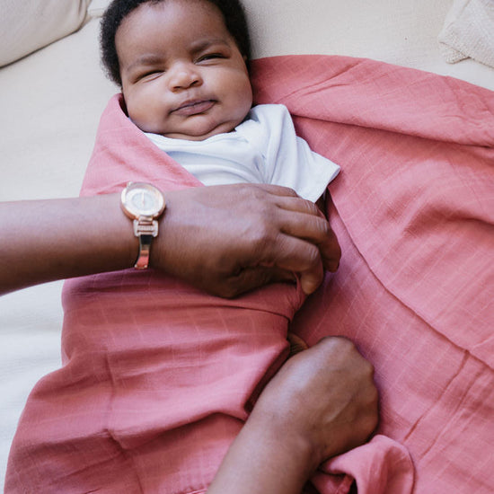 Child wrapped in a pink blanket with a hand holding their arm, lying on a couch.