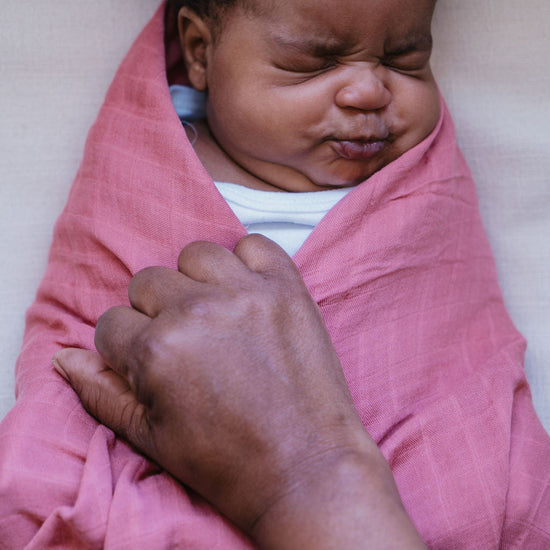 Newborn baby wrapped in a pink blanket with an adult hand holding onto it, on a light-colored surface.