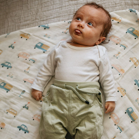 Baby lying on a patterned blanket wearing a white long-sleeve shirt and green pants.