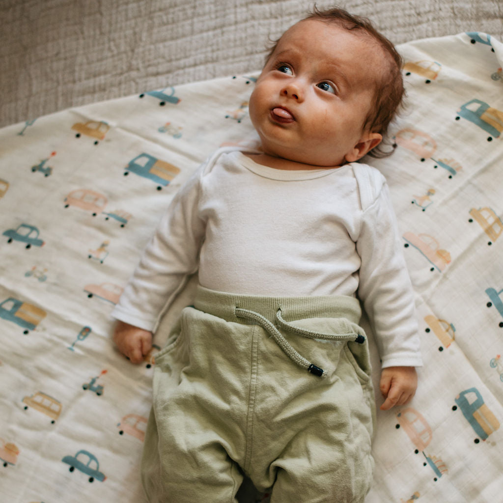 Baby lying on a patterned blanket wearing a white long-sleeve shirt and green pants.