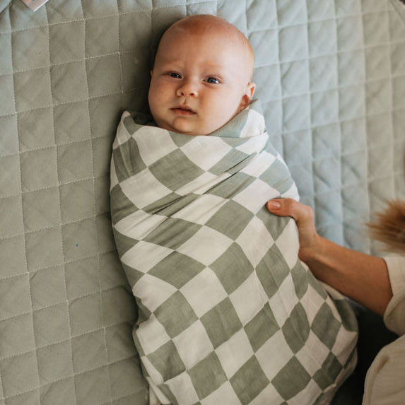 Baby swaddled in a green and white checkered blanket on a quilted mat with a person above.