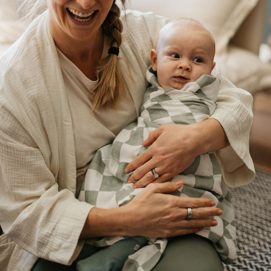 Woman holding a baby wrapped in a checkered blanket, both smiling indoors.