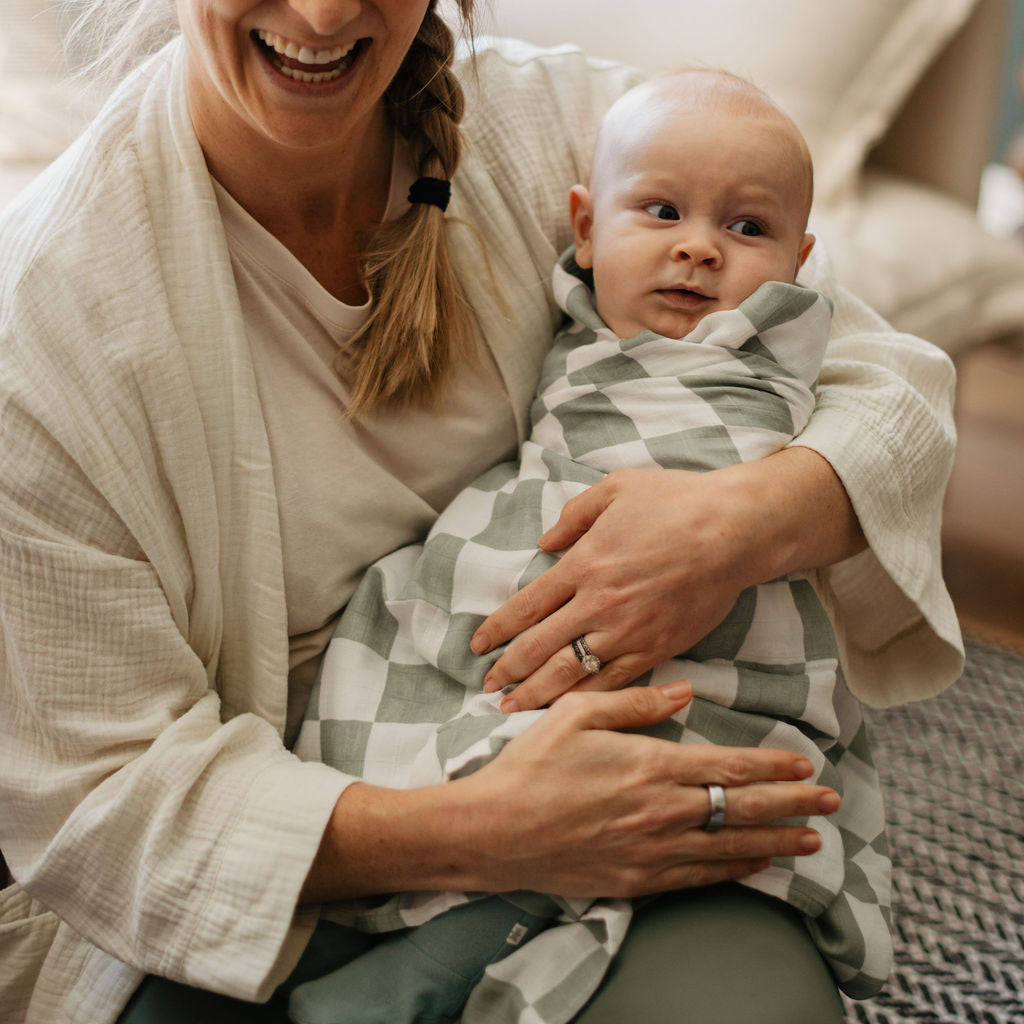 Woman holding a baby wrapped in a checkered blanket, both smiling indoors.