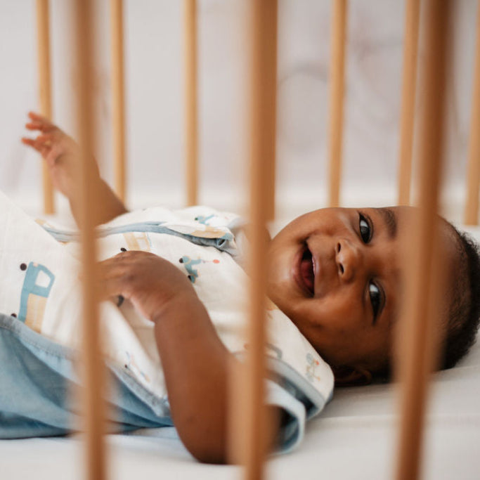 Baby lying in a crib with wooden bars, wearing a onesie with colorful letters.