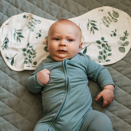 Baby in a green onesie lying on a textured surface with a decorative mat featuring leaf patterns.