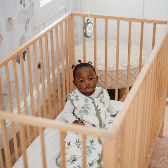 Child sitting in a crib wearing a white outfit with green patterns, surrounded by a neutral-colored room.