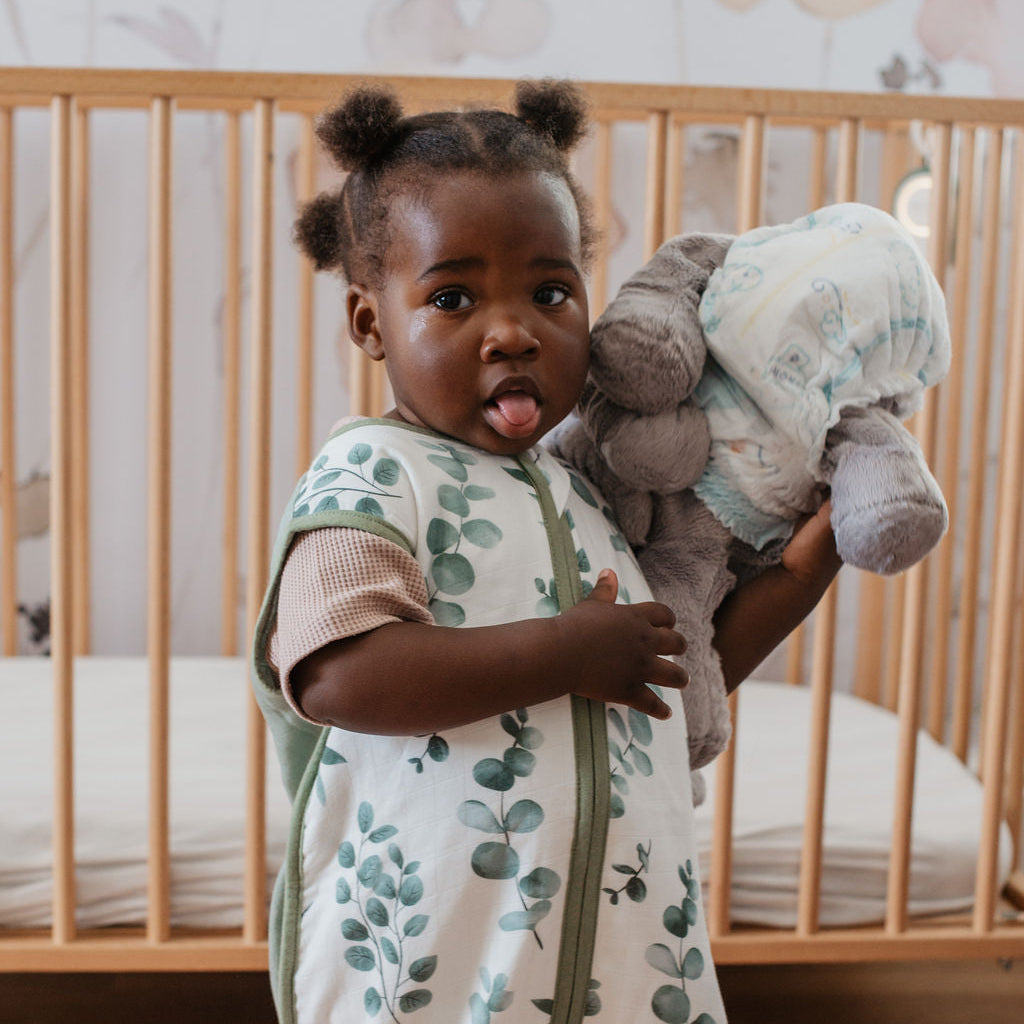 Child in a leaf-patterned swaddle holding a plush toy in a crib.