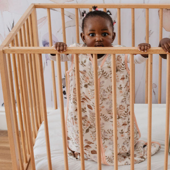 Two children in a crib with floral bedding, standing behind a wooden crib railing.