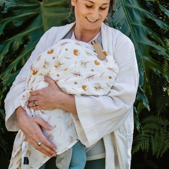 Woman holding a baby wrapped in a blanket with a natural pattern against a green leafy background