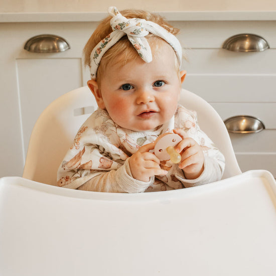 Baby sitting in a high chair wearing a floral outfit and headband, with a neutral background.