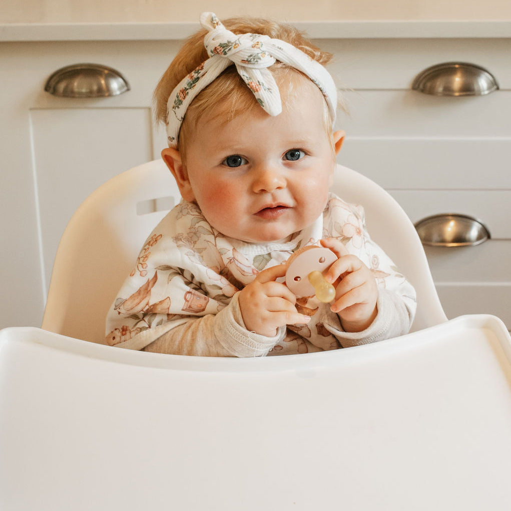 Baby sitting in a high chair wearing a floral outfit and headband, with a neutral background.