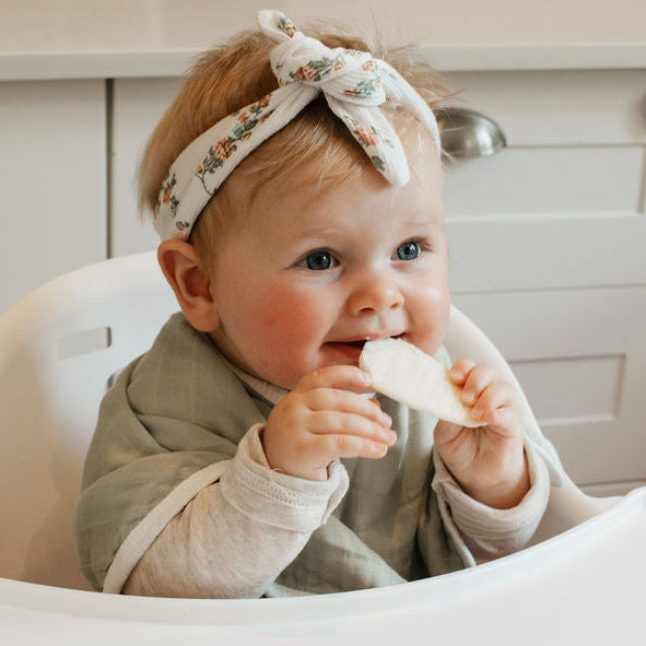 Baby sitting in a high chair with a bib, holding a piece of food.