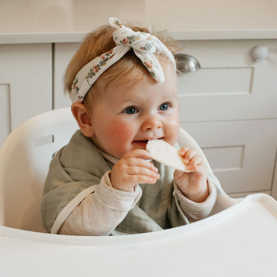 Baby sitting in a high chair with a bib, holding a piece of food.
