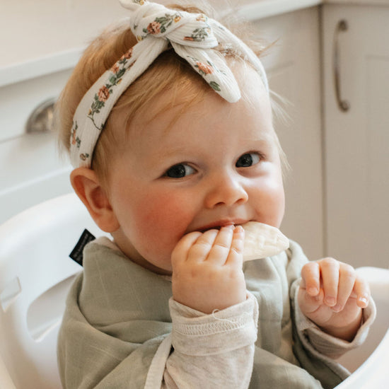 Baby sitting in a high chair wearing a floral headband and beige bib.
