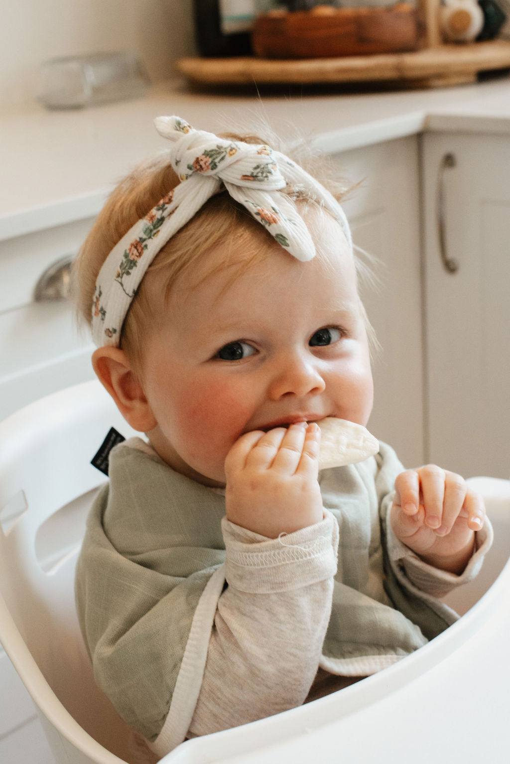 Baby sitting in a high chair wearing a floral headband and beige bib.