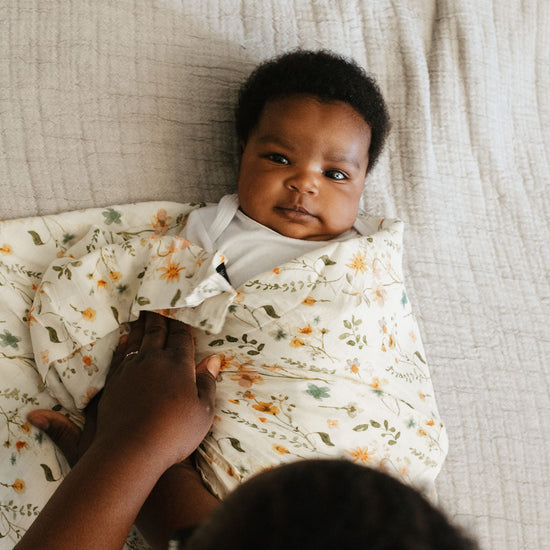 Baby wrapped in a floral blanket with an adult's hand, lying on a textured surface.
