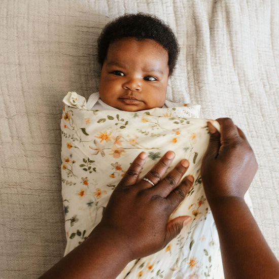 Baby wrapped in a floral blanket with adult hands holding it, on a neutral background